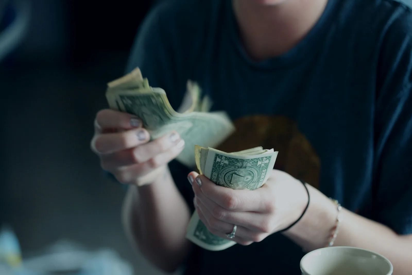 穿深色上衣的人雙手拿著一疊美元紙鈔正在清點，桌上有一個杯子